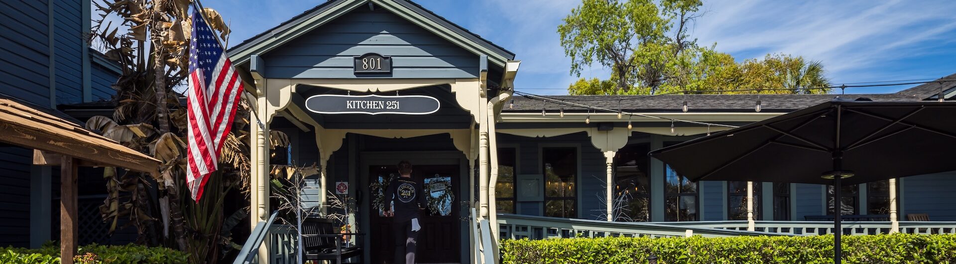 A small blue and gray restaurant with a covered porch, American flag, potted plants, and outdoor seating under a sunny blue sky with wispy clouds.