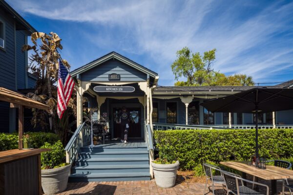 A small blue and gray restaurant with a covered porch, American flag, potted plants, and outdoor seating under a sunny blue sky with wispy clouds.