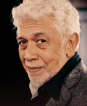 An older man with curly white hair, a trimmed white beard, and mustache, wearing a gray blazer over a black shirt, looks over his shoulder and smiles softly against a dark background.