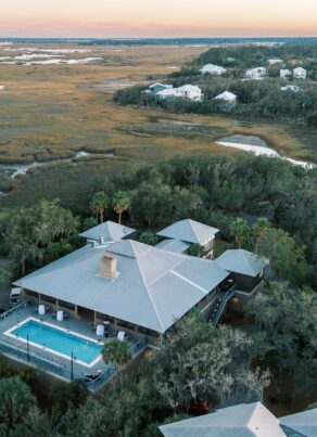 Aerial view of a house with a swimming pool surrounded by trees and marshland; nearby houses and winding waterways are visible in the background under a sunset sky.