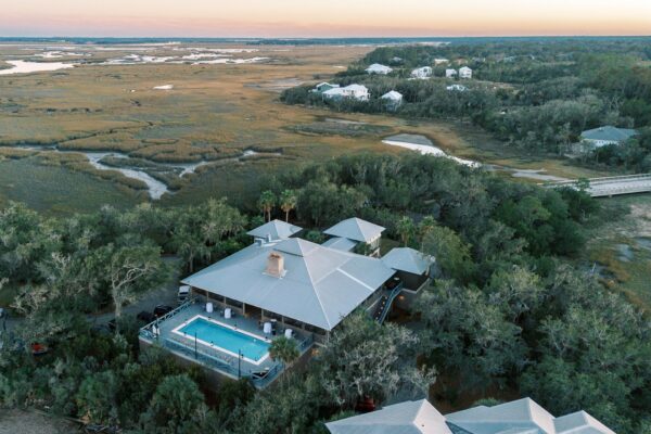 Aerial view of a house with a swimming pool surrounded by trees and marshland; nearby houses and winding waterways are visible in the background under a sunset sky.