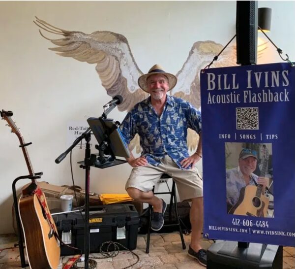 A smiling musician in a blue Hawaiian shirt and straw hat sits on a stool with instruments nearby. A sign reads Bill Ivins Acoustic Flashback with contact info and a photo of him holding a guitar. A mural of wings is on the wall behind him.