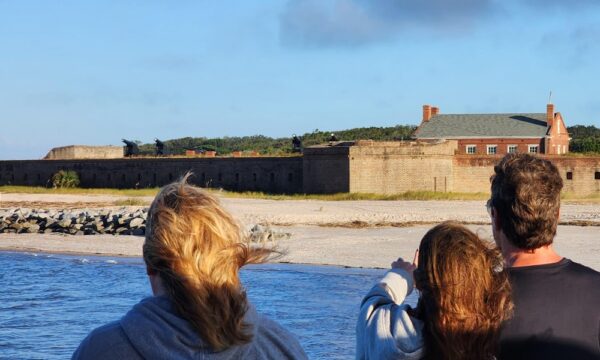 Three people stand near the water, looking toward a historic brick fort with cannons and a nearby building under a blue sky. The sandy shore and stone wall are visible in the background.