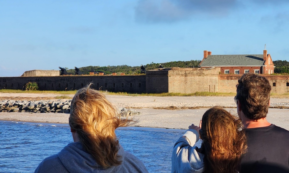 Three people stand near the water, looking toward a historic brick fort with cannons and a nearby building under a blue sky. The sandy shore and stone wall are visible in the background.