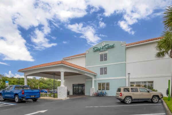 A hotel with a light blue and white exterior, red-tiled roof, and a covered entrance. Several cars and bicycles are parked outside. The sky is partly cloudy with palm trees visible.