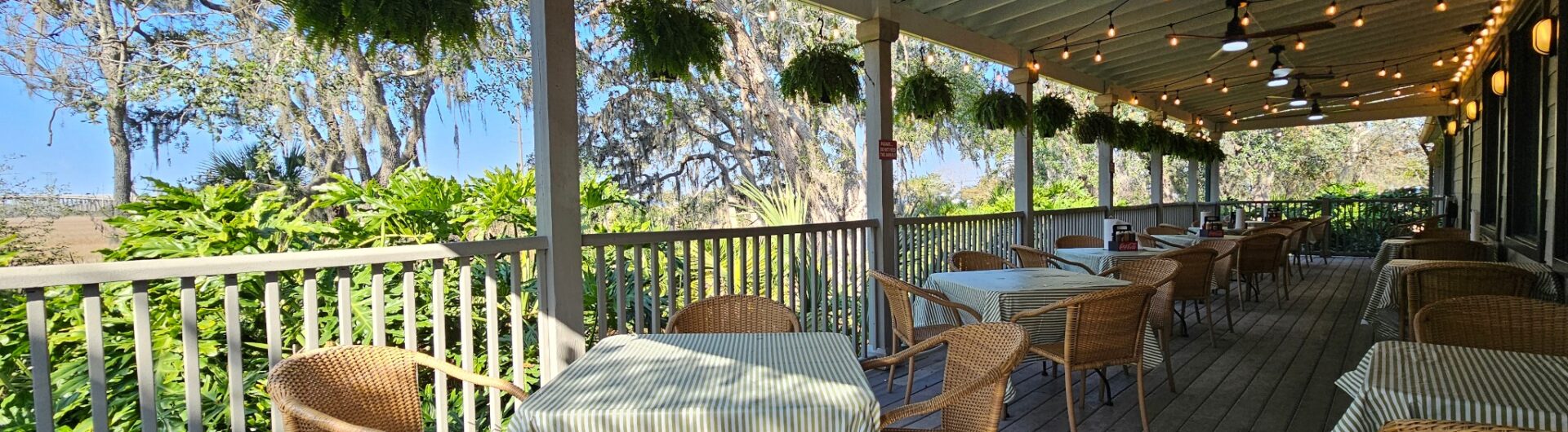 A covered outdoor patio with wicker chairs and tables, striped tablecloths, hanging plants, ceiling fans, and string lights, overlooking lush greenery and trees on a sunny day.
