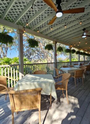 A covered outdoor patio with wicker chairs and tables, striped tablecloths, hanging plants, ceiling fans, and string lights, overlooking lush greenery and trees on a sunny day.