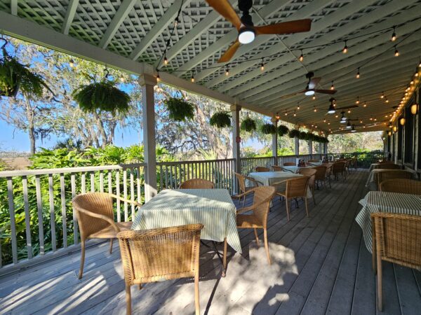 A covered outdoor patio with wicker chairs and tables, striped tablecloths, hanging plants, ceiling fans, and string lights, overlooking lush greenery and trees on a sunny day.