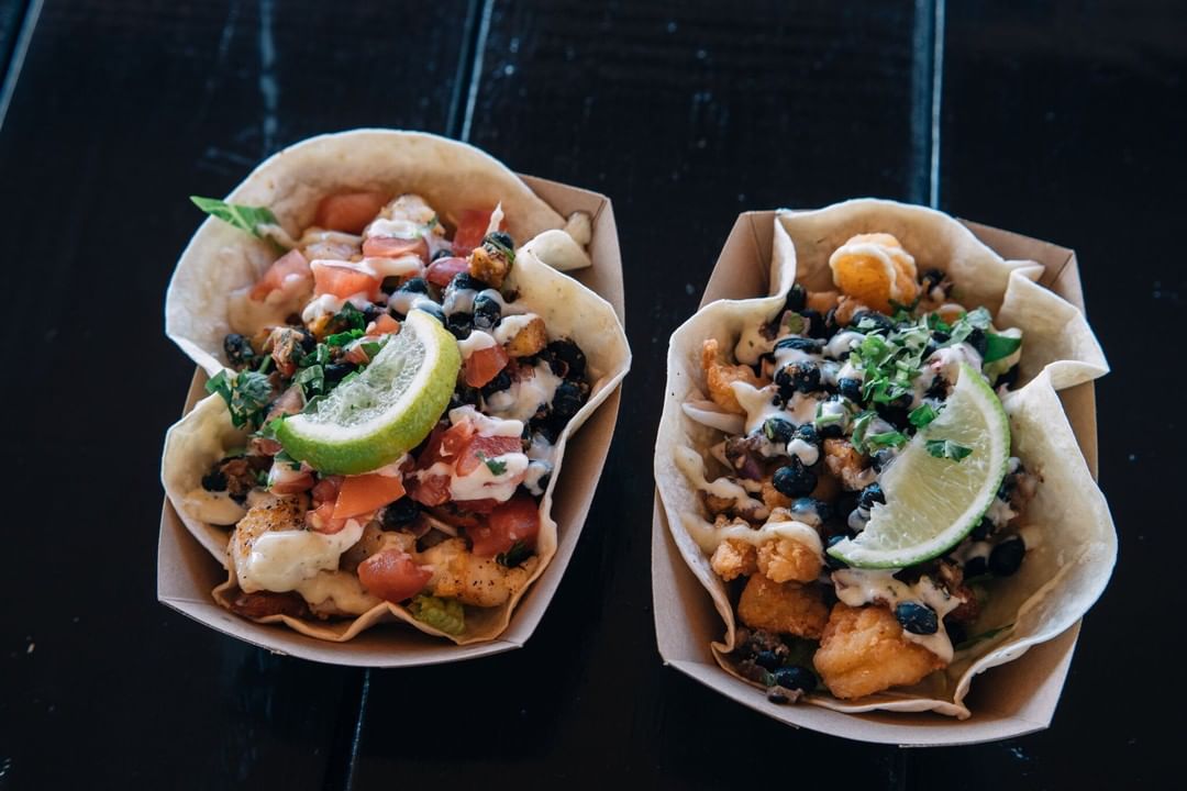 Two soft tacos in cardboard trays, topped with diced tomatoes, black beans, fried fish, chopped greens, creamy sauce, and lime wedges, served on a dark wooden surface.