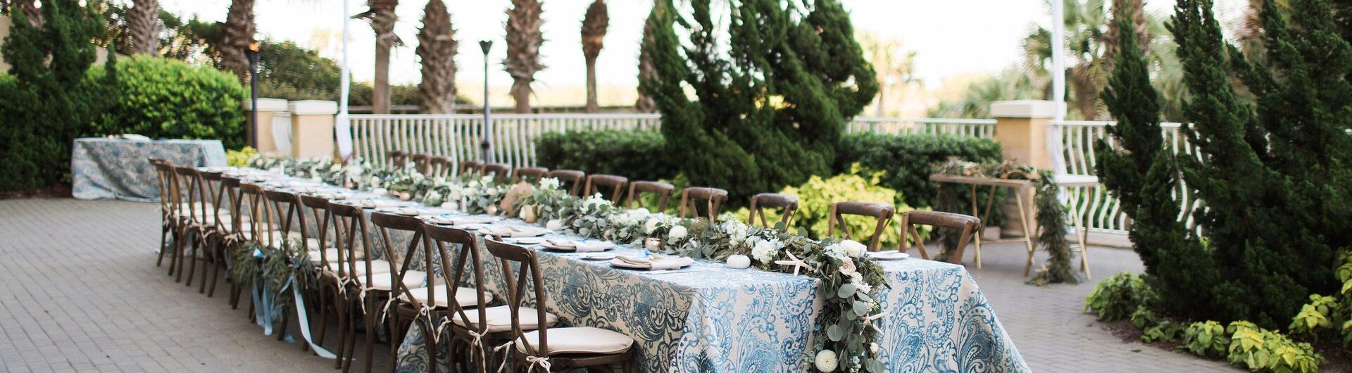A long outdoor table with patterned blue and white tablecloth, wooden chairs, and greenery garland is set on a brick patio with palm trees and string lights overhead.
