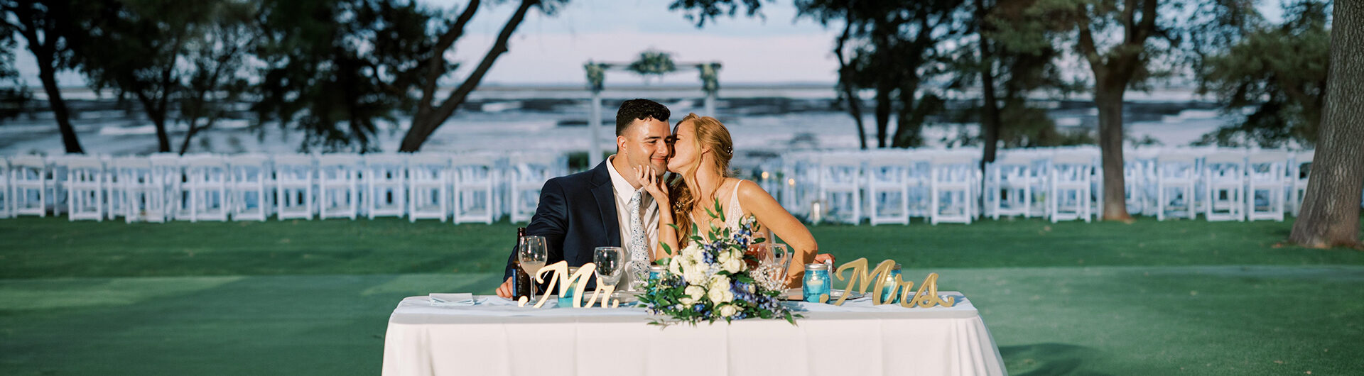 A bride and groom sit at a decorated table outdoors, with “Mr.” and “Mrs.” signs in front. The bride kisses the groom’s cheek as he smiles, and empty chairs and trees are visible in the background.
