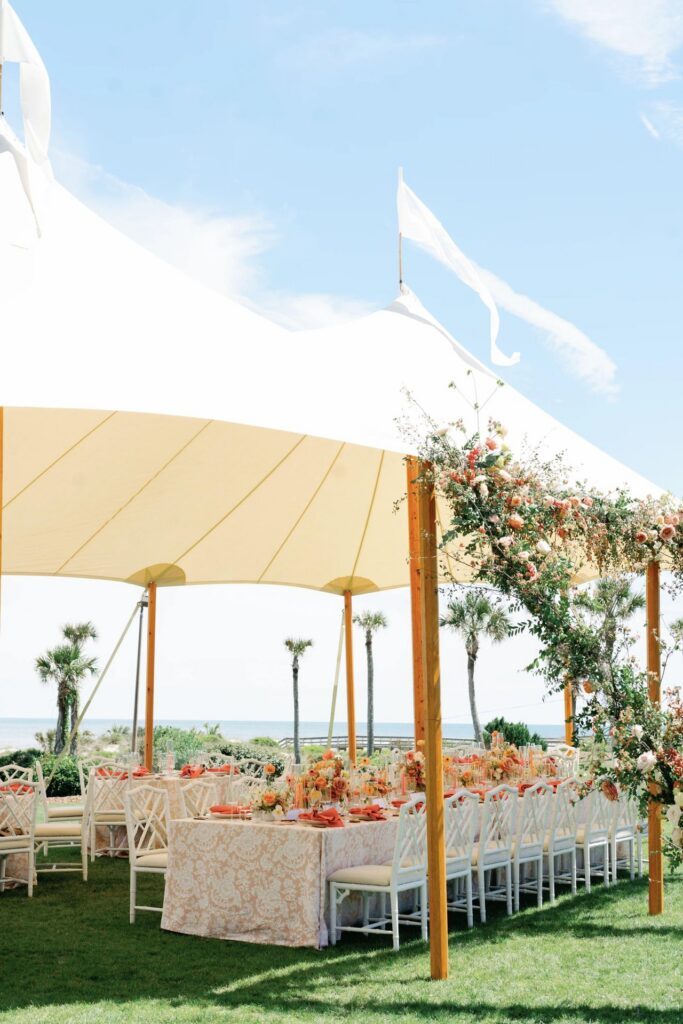 An outdoor wedding reception setup under a large white tent with long tables decorated with floral centerpieces, surrounded by white chairs, green grass, and palm trees with the ocean visible in the background.