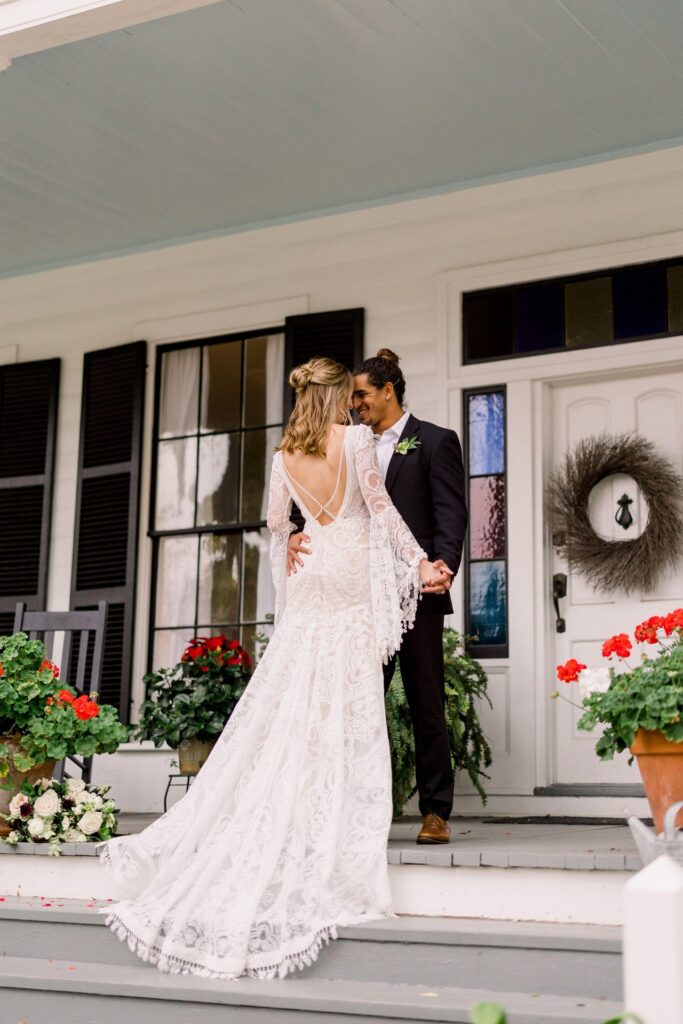 A bride in a long, lace white dress and a groom in a black suit stand holding hands on a porch decorated with potted flowers, gazing at each other and smiling.