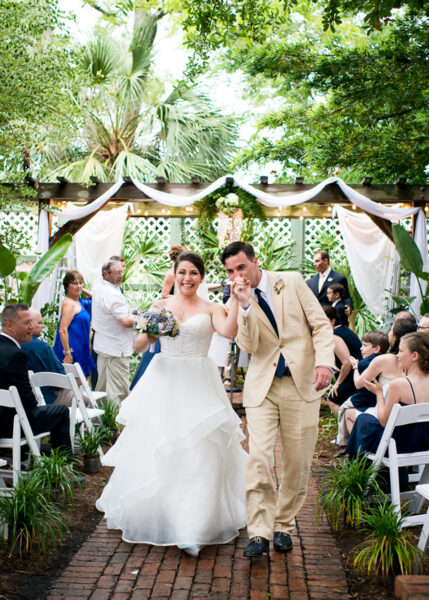 A bride in a white gown and a groom in a tan suit walk joyfully down an outdoor aisle, holding hands and smiling, surrounded by seated guests on white chairs and greenery.