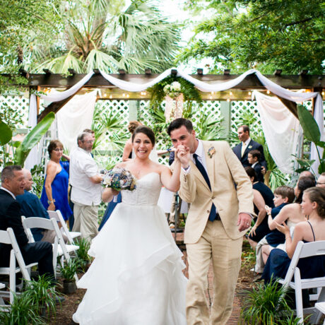A bride in a white gown and a groom in a tan suit walk joyfully down an outdoor aisle, holding hands and smiling, surrounded by seated guests on white chairs and greenery.